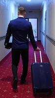 Man in suit with suitcase in hotel. Back view of a businessman with a trolley bag walking to his room in the hotel corridor. Vertical video
