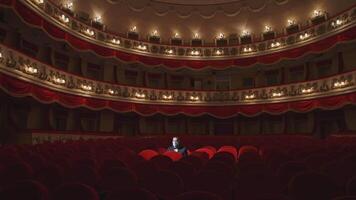 Spectator in mask sitting alone in empty theater. Man putting off his facial mask and talking in large dark auditorium among many rows of vacant red chairs in the theater. video