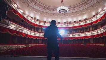 Actor on stage in spotlight. Active man standing on stage of a classical theatre with rows of red chairs without audience. Back view. video