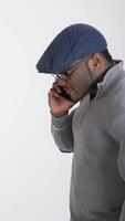 Serious black man with a phone in studio. Afro american guy in gray sweater and blue cap talking the mobile phone on white background. Side view. Vertical video