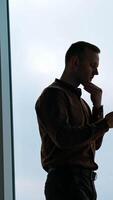 Young man on large window background. Businessman with a phone standing in office and looking through the window in the evening. Rear view. Vertical video