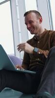 Smiling young man in modern office. Happy freelancer sitting on a floor and working on a laptop on window city view background. Vertical video