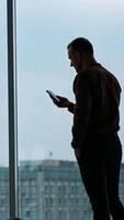 Office worker with a phone. Young businessman looking into the phone near the window in office on city buildings background. Panoramic window view. Vertical video