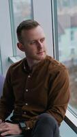 Thoughtful man sit on windowsill in office. Businessman with a laptop on laps looking at cityscape through panoramic window daydreaming about future. Vertical video