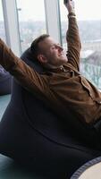 Relaxed businessman indoors. Happy young man is sitting by the window with stretched arms and having a rest in office on the city view window background. Vertical video