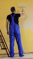 Room decoration with new color. Professional worker in blue overalls painting wall with a paint roller in empty room. Rear view. Vertical video