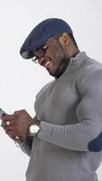 Stylish guy with black skin uses smartphone. Profile view of african man in cap and glasses typing messages on his mobile phone and smiling. Vertical video