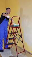 Young man in overalls renovating room in the house. Painter worker using a paint roller doing makeover of a room. Room decoration with new color. Vertical video