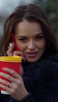 Attractive girl outdoors. Young woman in fur coat talking the phone while drinking coffee in the park. Portrait of a female with plastic cup. Slow motion. Vertical video