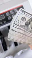 Bundle of one hundred dollar banknotes. Female's hands holding a large sum of money of dollar bills on the background of automatic counting machine. Vertical video