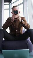Young man with a phone in office. Handsome businessman sitting on a comfortable armchair and using a mobile phone on window background. Front view. Vertical video