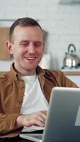 Young man freelancer using laptop in the kitchen. Happy man having a conversation through the laptop. Online working from home. Remote work from home during coronavirus quarantine. Vertical video