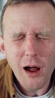 Portrait of a funny freelancer. Young man blinking eyes madly after sitting at home for a long period. Amusing man with a laptop on table in the kitchen. Lockdown concept. Vertical video