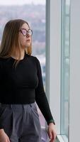Confident business woman in modern office. Beautiful girl in glasses and stylish clothes standing near the large office windows and looking on the city view. Vertical video