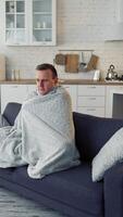 Man having fever. Young man feeling freezy and covering with warm blanket while sitting on a couch at home. Sick man wrapped with blanket during covid outbreak. Vertical video
