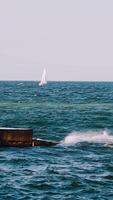 Motion of sea water. Waves crashing on stone harbor making white foam. White sail floating on the water surface. Slow motion. Vertical video