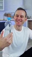 Smiling young man with a bottle of water at home. Young man proposing fresh water after the workout and looking on camera indoors. Vertical video