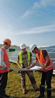 Male technicians with a project plan on a solar farm. Three workers in protective uniform and helmets discussing about solar panels installation in a bright day. Vertical video