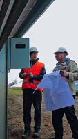 Solar technicians install photovoltaic panel on the field. Two engineers in protective uniform and hard hat with project plan under the big solar panel. Vertical video