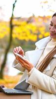 Happy smiling young woman using gadgets resting in the nature. Lady looks at her phone smiling and having laptop on her laps. Blurred nature autumn backdrop. Vertical video