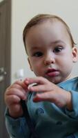 Baby boy in blue shirt sits on the floor looking curiously on the shoe lace in his hands. Cute toddler plays with a cord waving it cheerfully. Low angle view close up. Vertical video