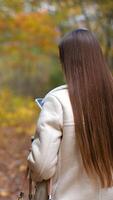 Long-haired woman walks looking at her phone with a paper cup in her hand her back to camera. Smiling lady turns around and makes few steps backwards. Autumn park at backdrop. video