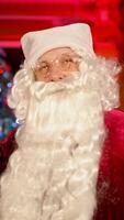 Santa Claus smiling on camera indoors. Portrait of a real Santa with white beard, in glasses and red costume standing in the living room on Christmas tree background. Vertical video