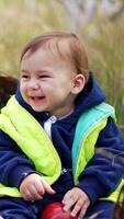 Beautiful smiling kid sitting on the grass with apples around. Adorable kid laughs sweetly and looks at the red apple with interest. Blurred nature at backdrop. Vertical video