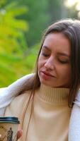 Good-looking Caucasian lady enjoying the vibes of warm autumn nature. Brunette sits on the ground with a paper cup in her hand. video