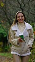 Happy smiling brunette long-haired lady walks with paper cup in the park. Lady smiles to the camera gesturing calling with her hand. Autumn park backdrop. video