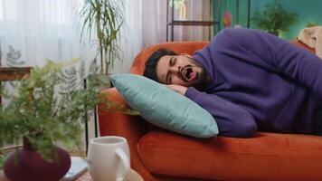 Tired indian man lying down in bed taking a rest at home napping falling asleep on sofa relaxing video