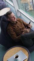 Young businessman sitting at break time. Smiling man sitting by the window and looking into the phone on the city background through the window. Top view. Vertical video