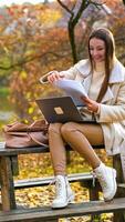 Happy woman working remotely in the autumn park. Lady checking up the documents and holding computer on her laps. Blurred backdrop. Vertical video