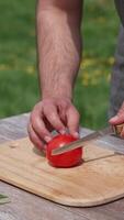 Man cutting tomato on a board outdoors. Preparing dinner at dacha. Man cuts vegetable into four parts on a table before the picnic. Vertical video