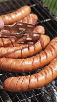 Roast wiener on a barbecue grill on charcoal. Cook turns over hot grilled sausages with a food tong in the garden in summer. Delicious food for a picnic. Vertical video