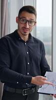 Surprised man working in office pointing at chart in his hands. Businessman surprised with results shown in graphs. Vertical video