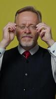 Funny grey-haired man putting on glasses. Senior man in elegant clothing with eyeglasses making amusing eyes on yellow background. Slow motion. Vertical video
