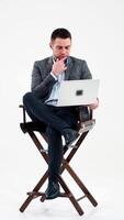 Thoughtful businessman with laptop isolated on white. Handsome man in elegant costume sitting on a high stool and working on the laptop. Business concept. Vertical video