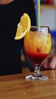 Tasty cocktail in glass. Bartender cooks the beverage for client. Fresh alcohol drink ready on a bar table. Slice of orange on a glass with color liquid with ice. Vertical video