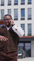 Waist up portrait view of the handsome businessman is having friendly call with his family, friends or partners. Young man is walking outside on business district on sunny warmly day Vertical video