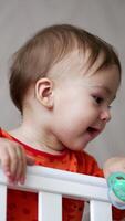 Beautiful Caucasian baby boy stays up in white cot. Cute kid playing with pacifier throwing it on the floor and watching it fall. Low angle view. Vertical video
