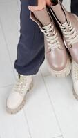 Female model wearing loose pants and white tractor sole boots holding another pair of beige boots in hands. View from above. White floor backdrop. Vertical video