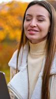 Beautiful smiling lady holding a laptop speaking to the camera. Dark-haired woman spending time in the nature in autumn. Vertical video
