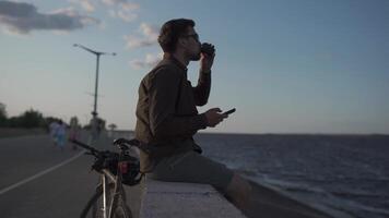 Man cyclist resting overlooking sea, drinking coffee and using smartphone while sitting on embankment. Male 30s wearing sunglasses, drinking takeaway coffee and using mobile phone on waterfront. video