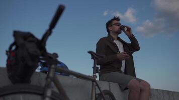 Traveler on bicycle stopped to enjoy the view and check road map on smartphone to the sea and have coffee in cycle time of travel. Male cyclist use smartphone and drinks takeaway coffee on embankment. video