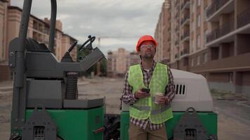 Construction worker on high-rise residential building site takes break, wearing hard hat and reflective vest. Relaxing near heavy equipment, enjoys coffee and uses smartphone before returning to work. video