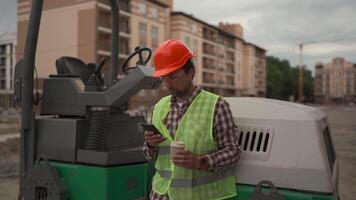 Construction worker stands at a high-rise building site, taking a break with a coffee and checking his smartphone. Dressed in a hard hat and reflective vest, he stands near heavy machinery and tools. video