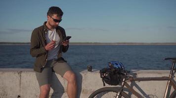 Bicycle rider eats lunch and surfs web on smart phone in front of pond. Man eating and using his phone while cycling before lake. Having lunch on a bicycle outdoors. Snack during commuting by bike. video
