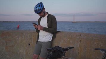 Theme is cycling in seaside. Male cyclist stopped to enjoy the sea view and see the road on the map on his smartphone. A male with a bicycle on the embankment is resting and using a smart phone. video