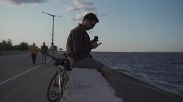 Traveler on bicycle stopped to enjoy the view and check road map on smartphone to the sea and have coffee in cycle time of travel. Male cyclist use smartphone and drinks takeaway coffee on embankment. video